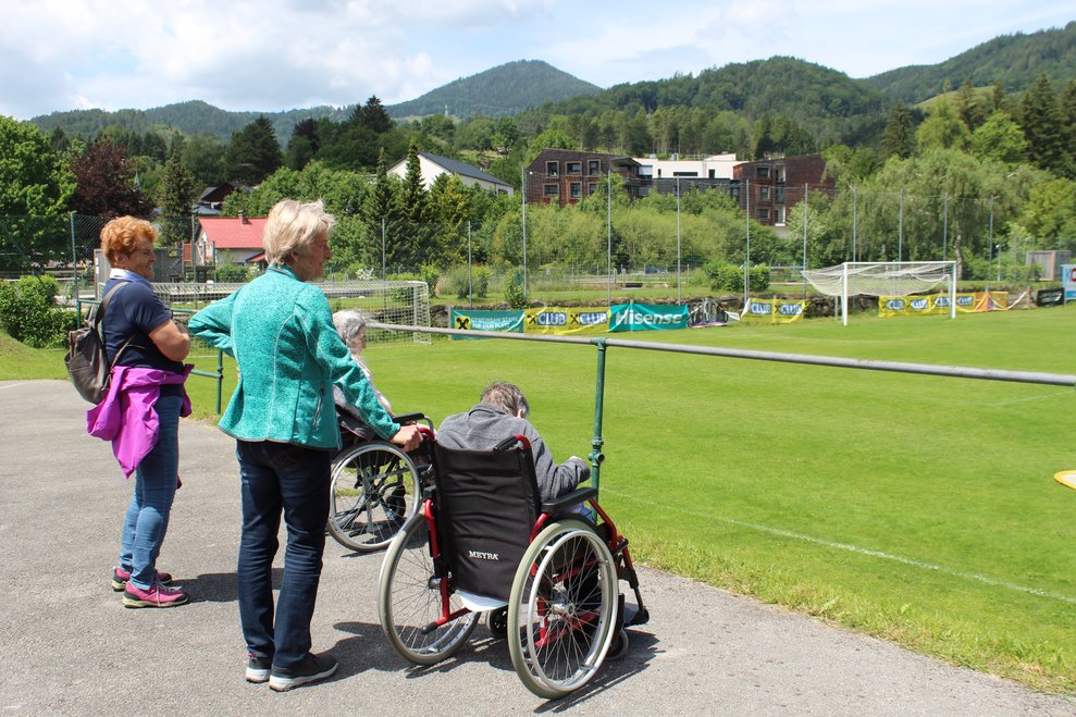Ehrenamtliche Mitarbeiterinnen und Bewohnerinnen im Rollstuhl feuern vom Spielfeldrand aus die Spielenden an